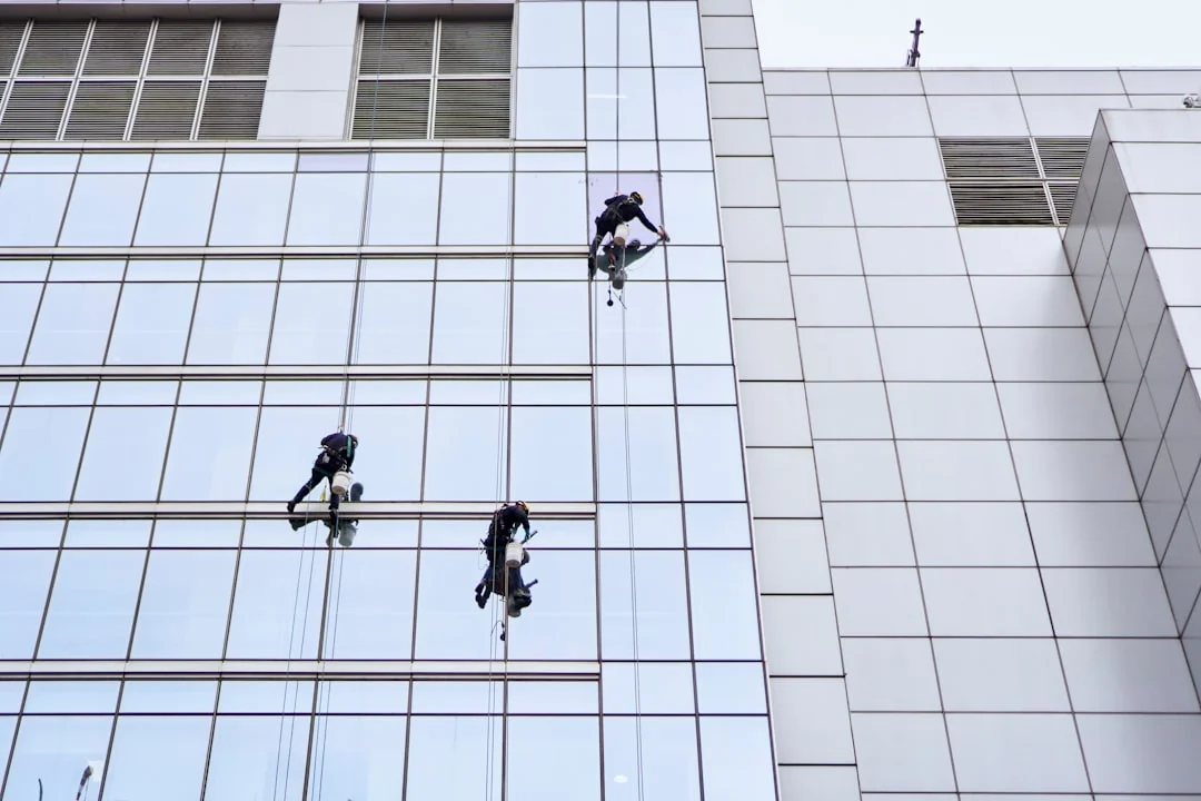 services-03 Two professional window cleaners working on a tall glass building using suspended harnesses.